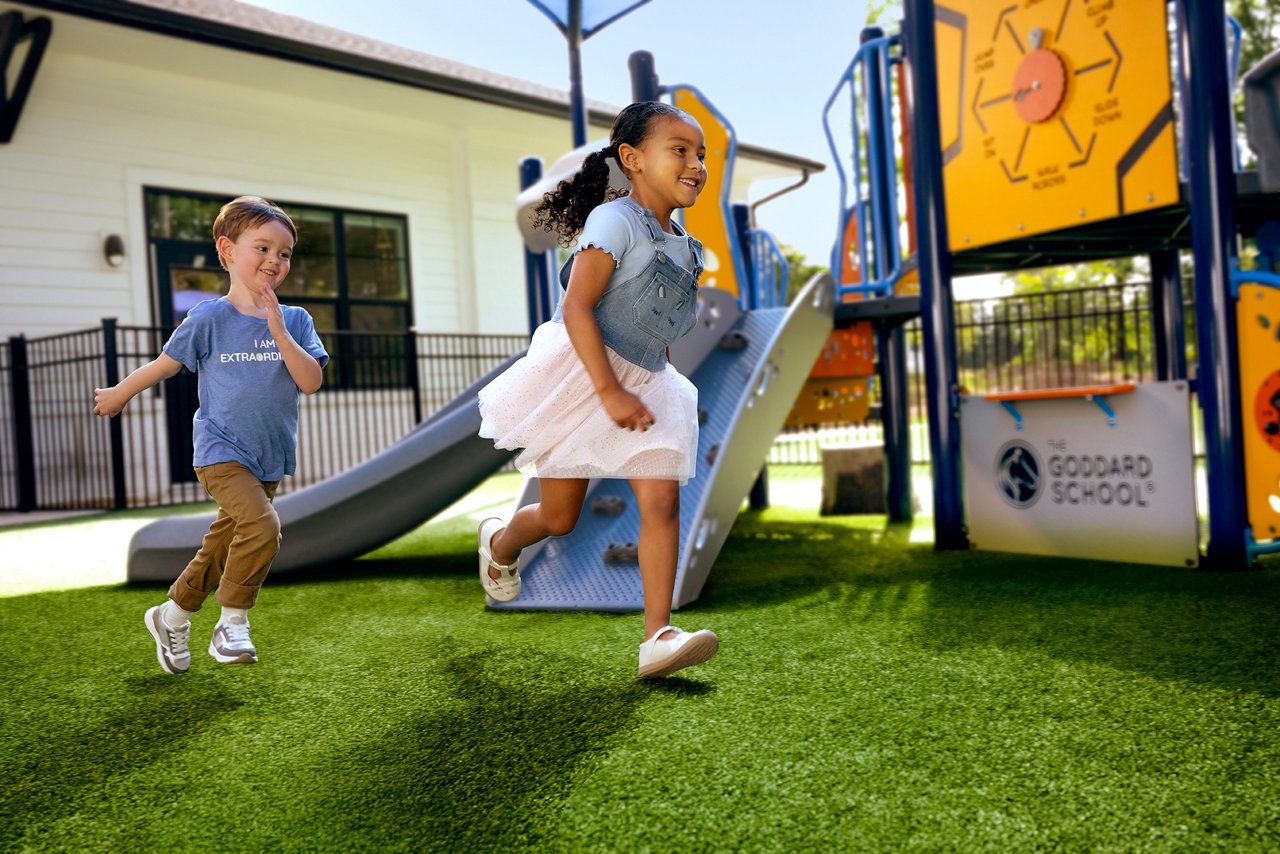 Two children running on artificial grass at a playground outside The Goddard School, with a slide and colorful play equipment in the background.