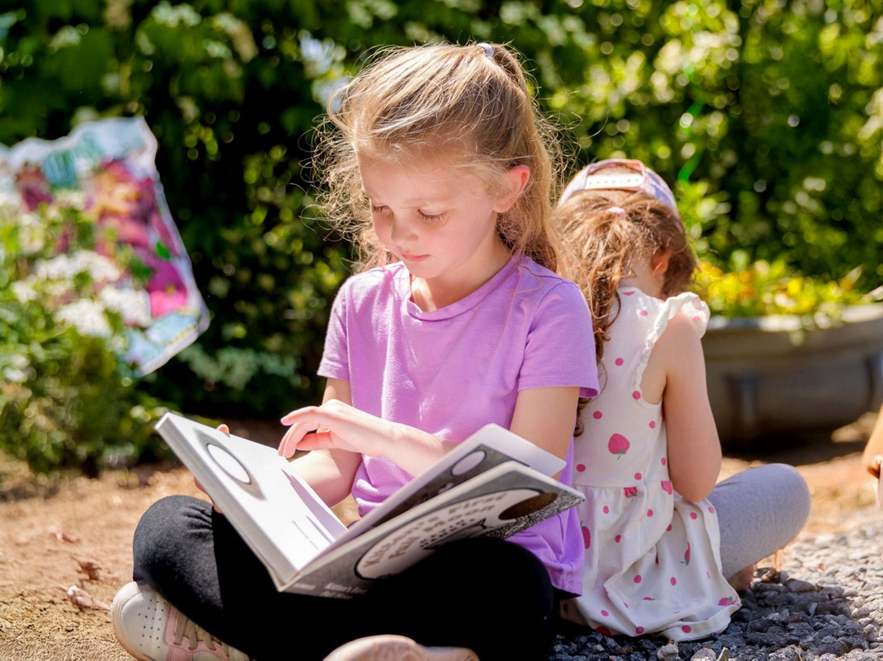 Preschool children reading outdoors surrounded by greenery and colorful artwork, promoting literacy and nature-based learning in an engaging early education setting.