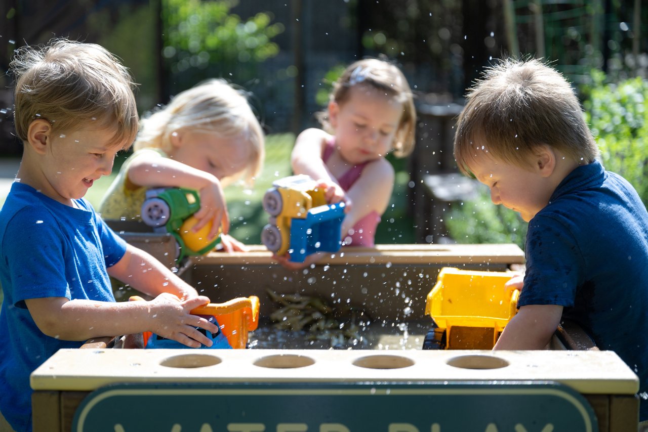 Preschool children enjoying sensory water play with scooping and pouring toys at a labeled water table, fostering early STEM skills and social development.
