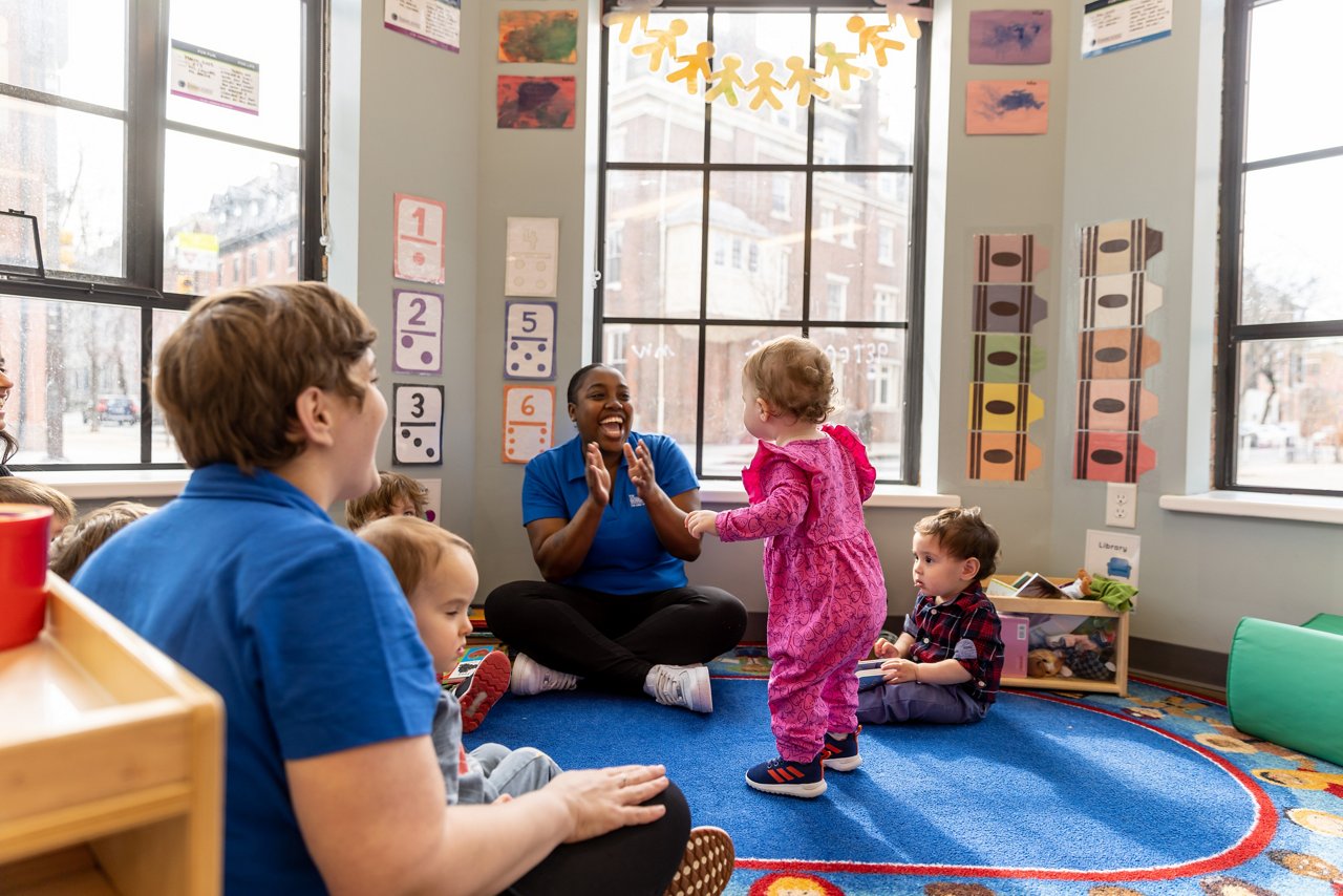 Group of preschool children and teachers participating in a classroom activity on a blue circular rug, surrounded by educational posters and natural light—promoting early learning, social interaction, and a joyful preschool environment.