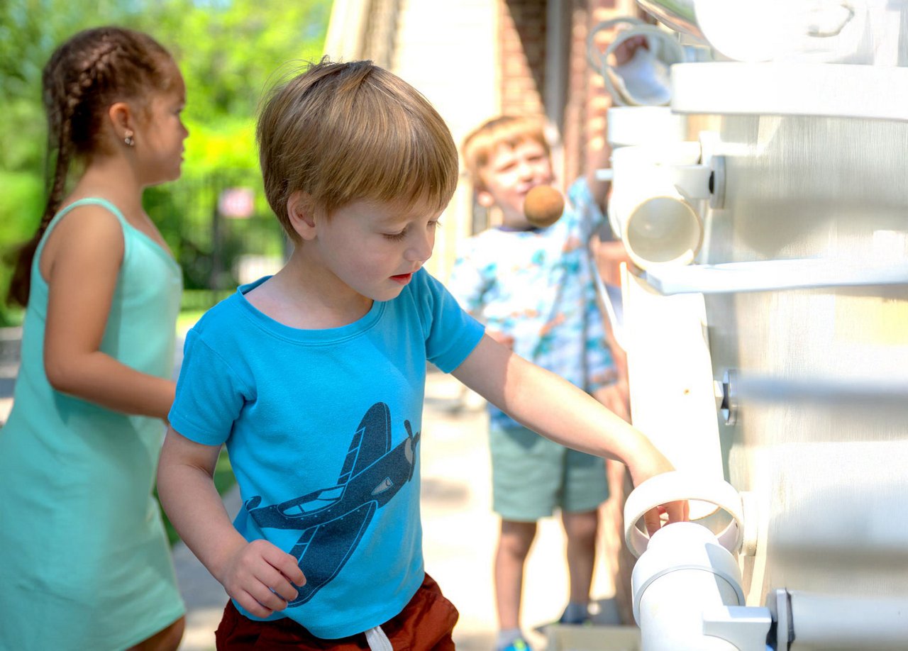 Preschool children exploring an outdoor play structure with tubes and openings, encouraging hands-on discovery, problem-solving, and collaborative play in a nature-based learning environment.