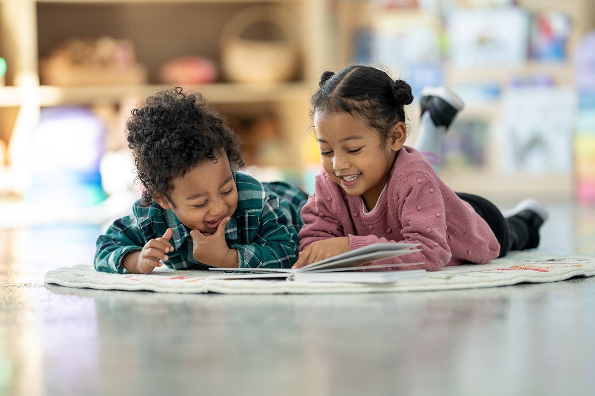Two preschool children lying on a classroom rug and reading a picture book together in a bright, organized learning space.