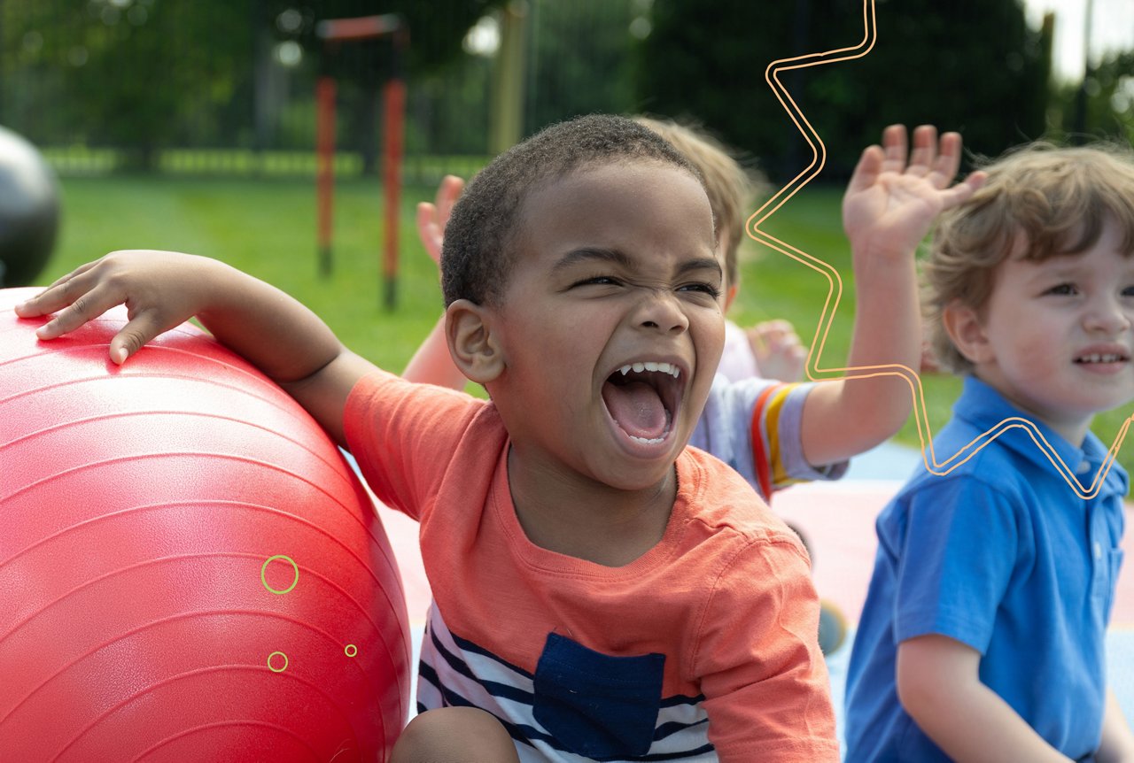 A young boy with a big smile holding a large red ball outside on a preschool playground with other children