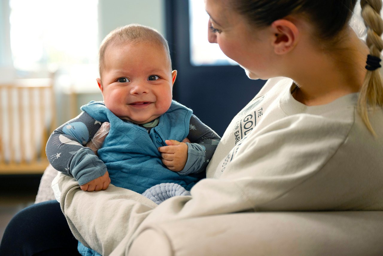 A smiling baby in a blue outfit sits on a teacher's lap. The woman, with a braided hairstyle, looks at the baby affectionately indoors. Bright and cheerful.