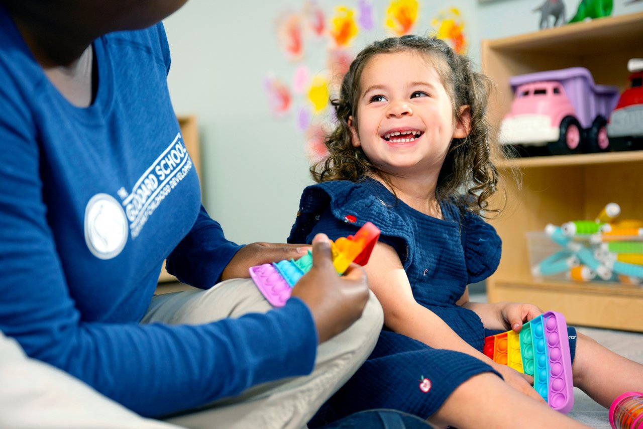 A young girl in a blue dress smiles joyfully while holding colorful sensory toys. She sits beside an adult in a playroom with shelves of toys and a bright, cheerful atmosphere.
