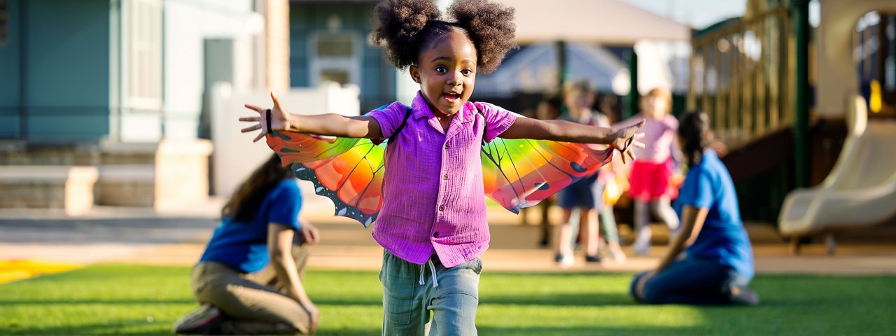 A child wearing colorful butterfly wings holding her arms open, smiling and running around a preschool playground