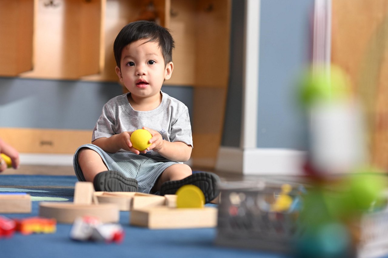 A young child sits on a blue carpet in a playroom, holding a yellow ball. Wooden blocks are scattered around, creating a playful and joyful atmosphere.