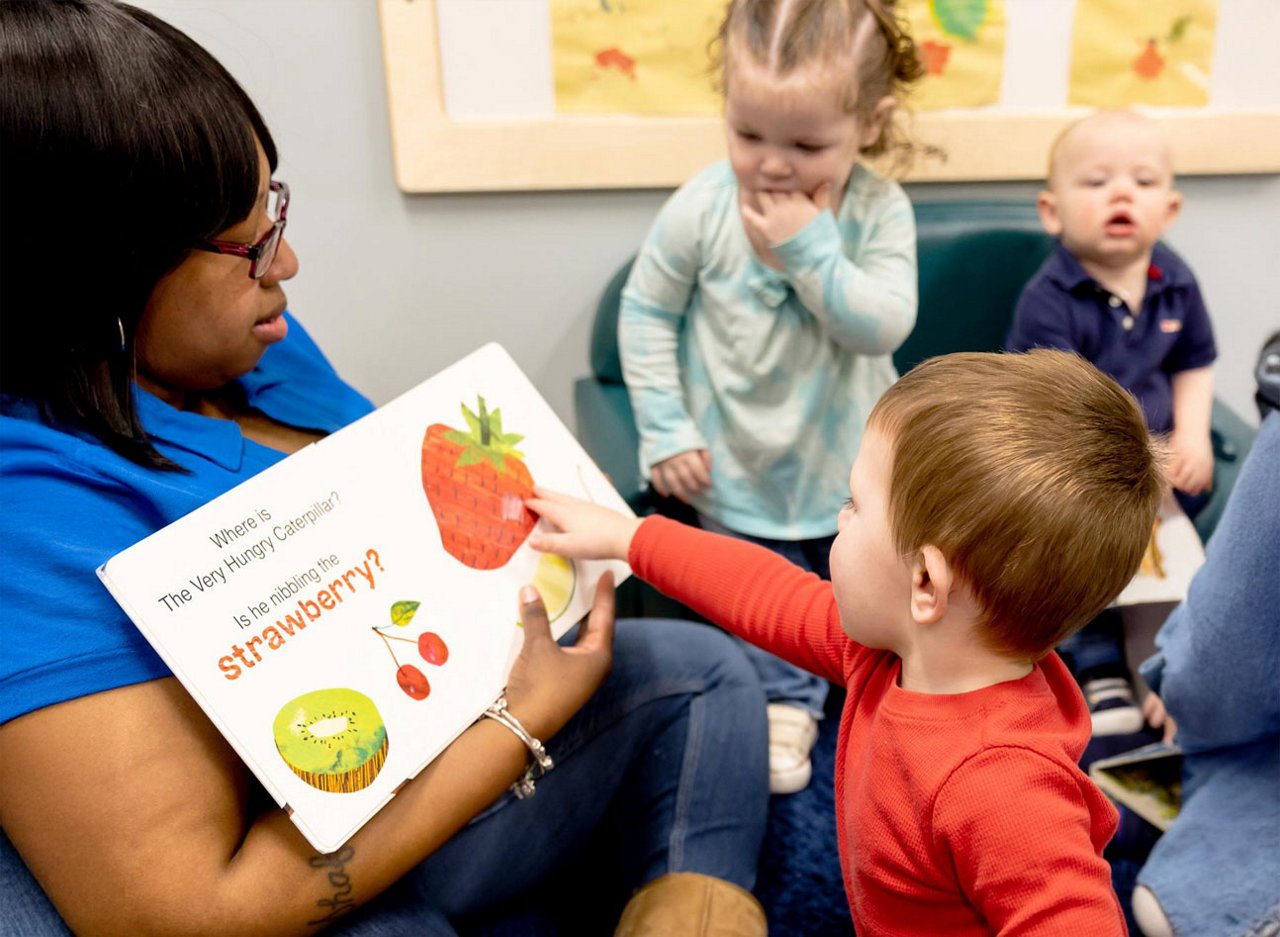 Preschool teacher reading a fruit-themed book with children in a classroom, encouraging early literacy, curiosity, and interactive storytime in a nurturing learning environment.
