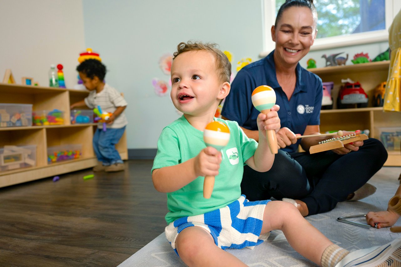 A toddler joyfully shakes maracas while sitting on the floor, supervised by a smiling adult. Another child plays nearby. The room is bright and colorful.