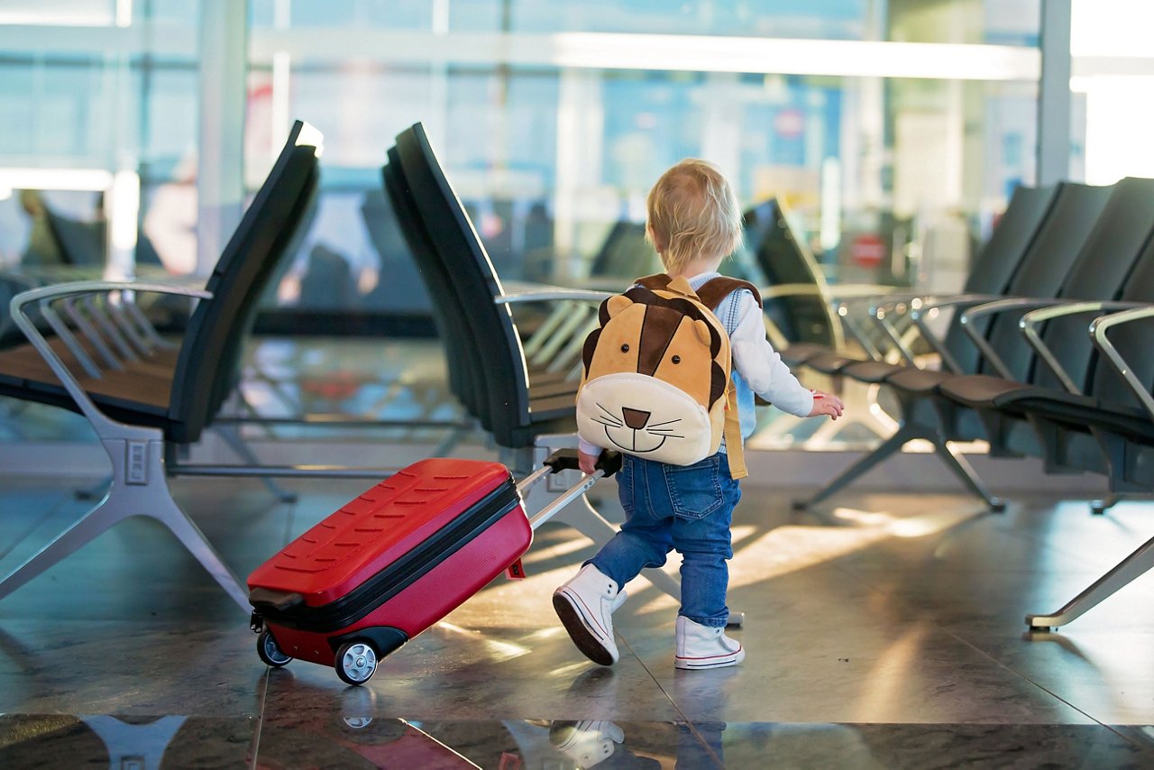 A young child wearing a lion backpack and pulling a red suitcase through an airport seating area.