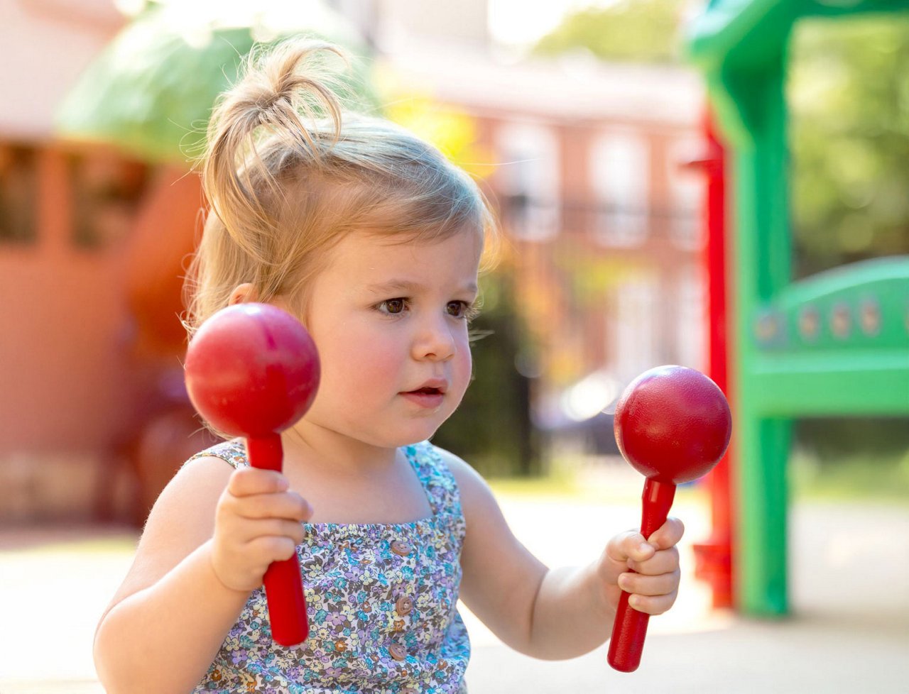 Preschool child playing with red maracas in an outdoor playground — music and movement activity supporting rhythm, coordination, and early childhood sensory development.