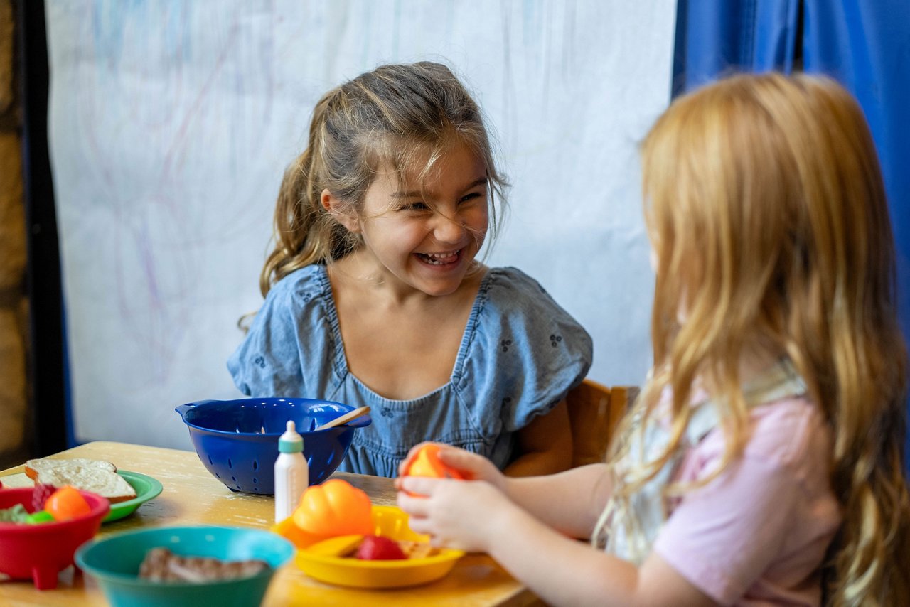Two young girls laughing and chatting while playing with toy food at a table in a preschool classroom