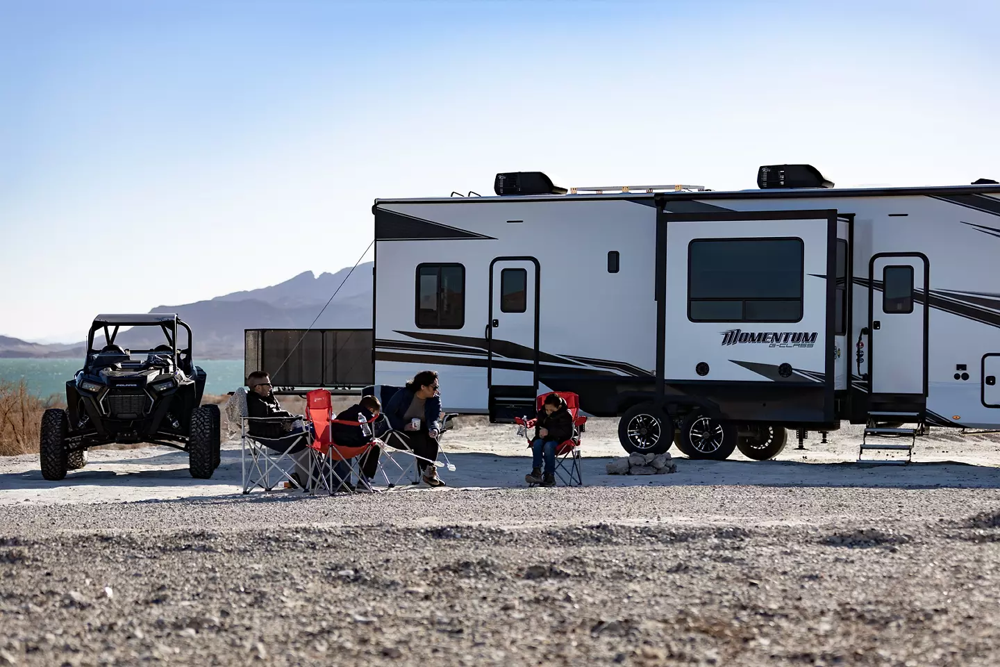 Polaris next to Momentum Toy Hauler, with campers sitting in camping chairs in front of the RV. 