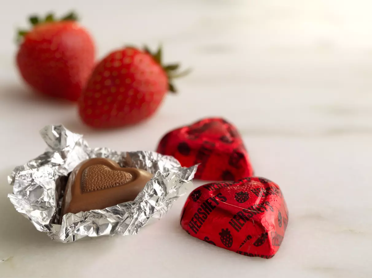 Wrapped and unwrapped HERSHEY'S Strawberry Creme Hearts beside pair of Strawberries