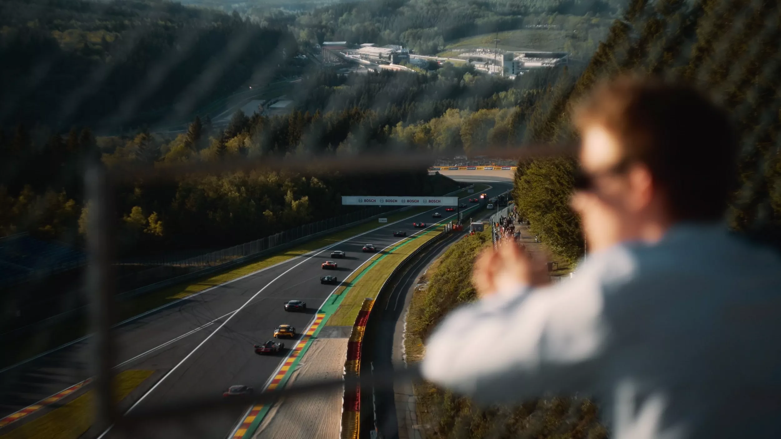 Race cars on a winding forest track, with a cheering spectator blurred in the foreground