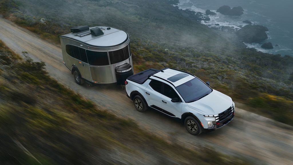White Hyundai Santa Cruz towing a small silver travel trailer on a gravel road above the ocean