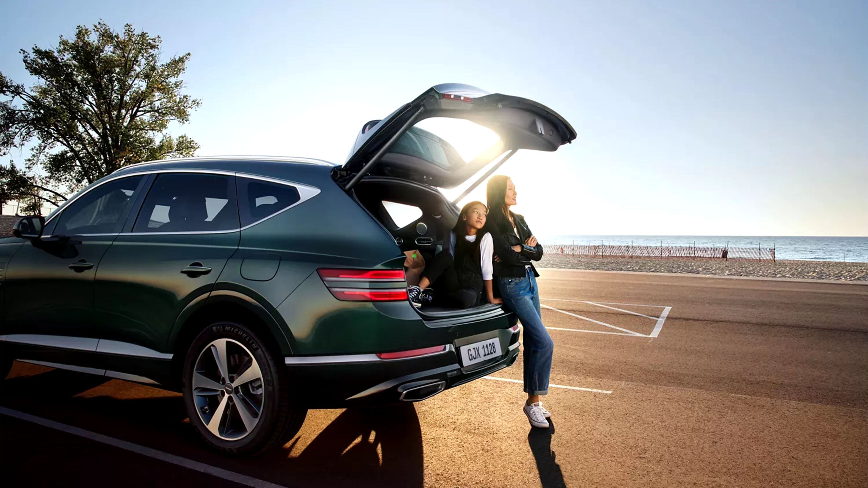 Green Genesis SUV parked by a beach with its liftgate open, a woman and a child resting in the cargo area at sunset