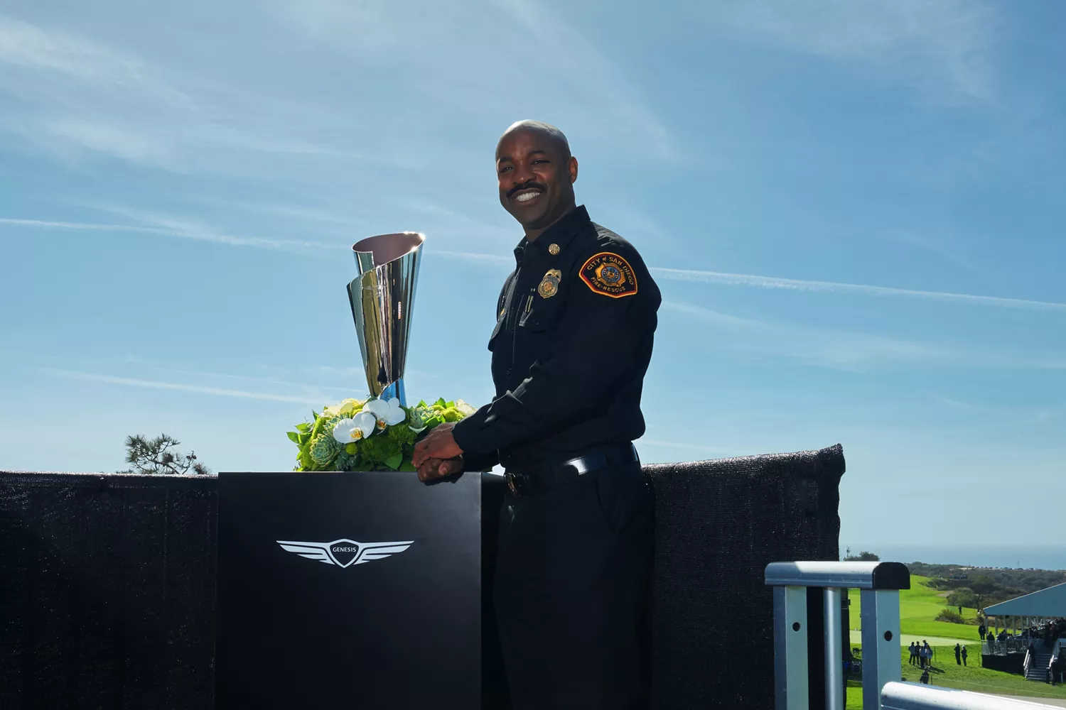 Firefighter standing next to Genesis Invitational trophy at Genesis Invitational, fairway in back