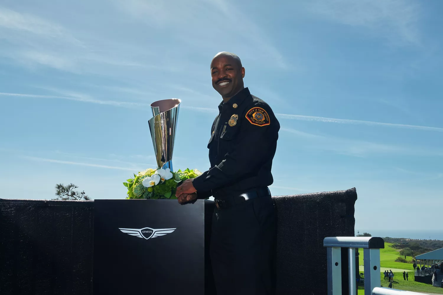 Firefighter standing next to Genesis Invitational trophy at Genesis Invitational, fairway in back