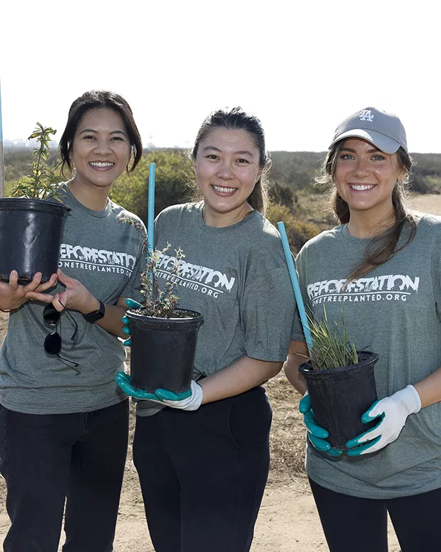 Two women and one man in matching teal t-shirts and white hats plant a tree during a Hyundai event