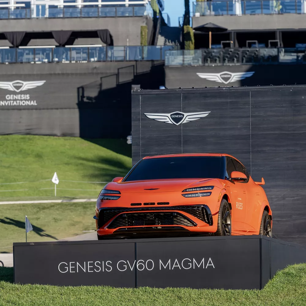 Front driver side view of orange Genesis GV60 Magma on display platform at 2026 Genesis Invitational