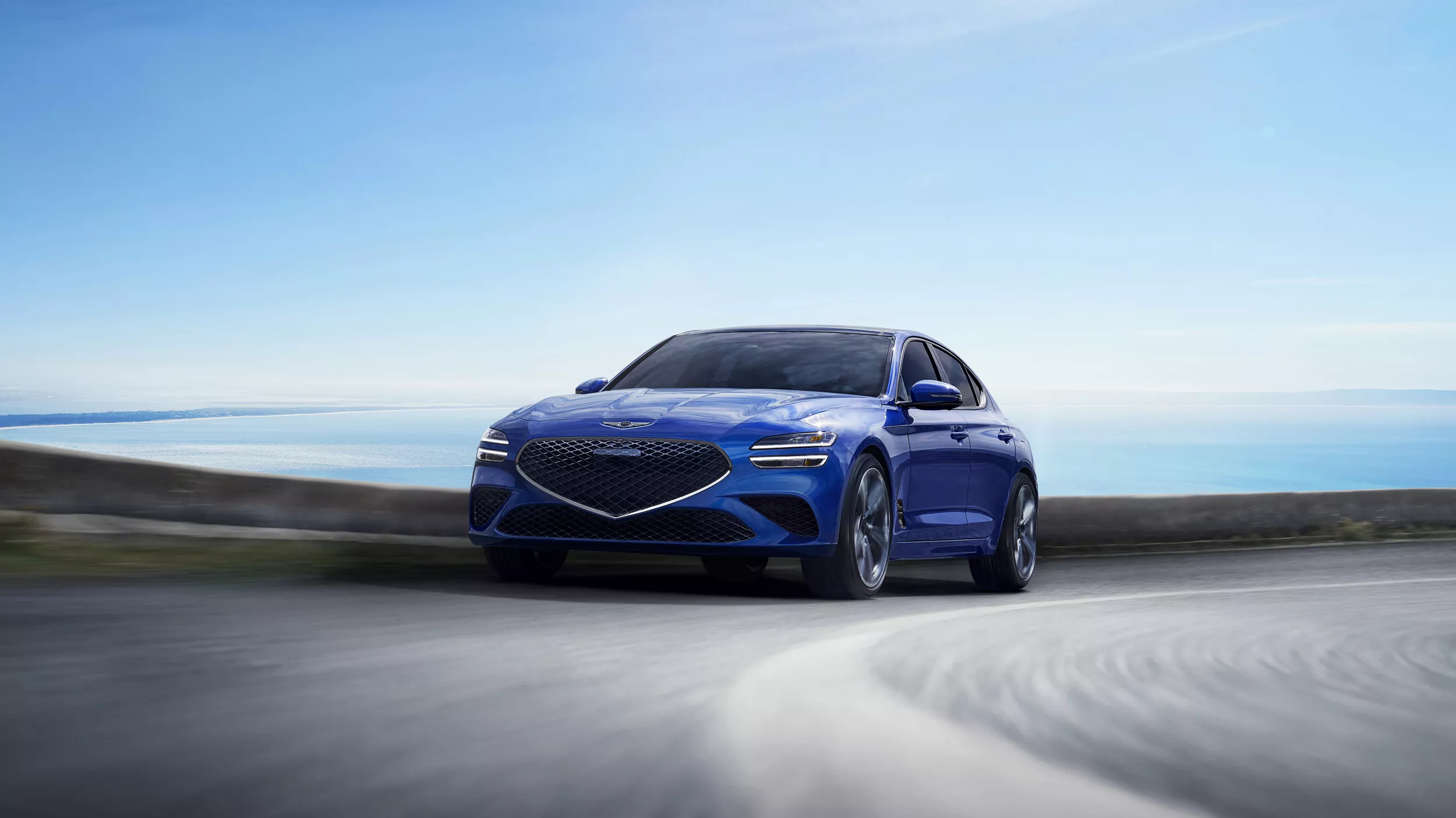 Front view of blue 2025 Genesis G70 rounding a corner with the ocean and sky in the background