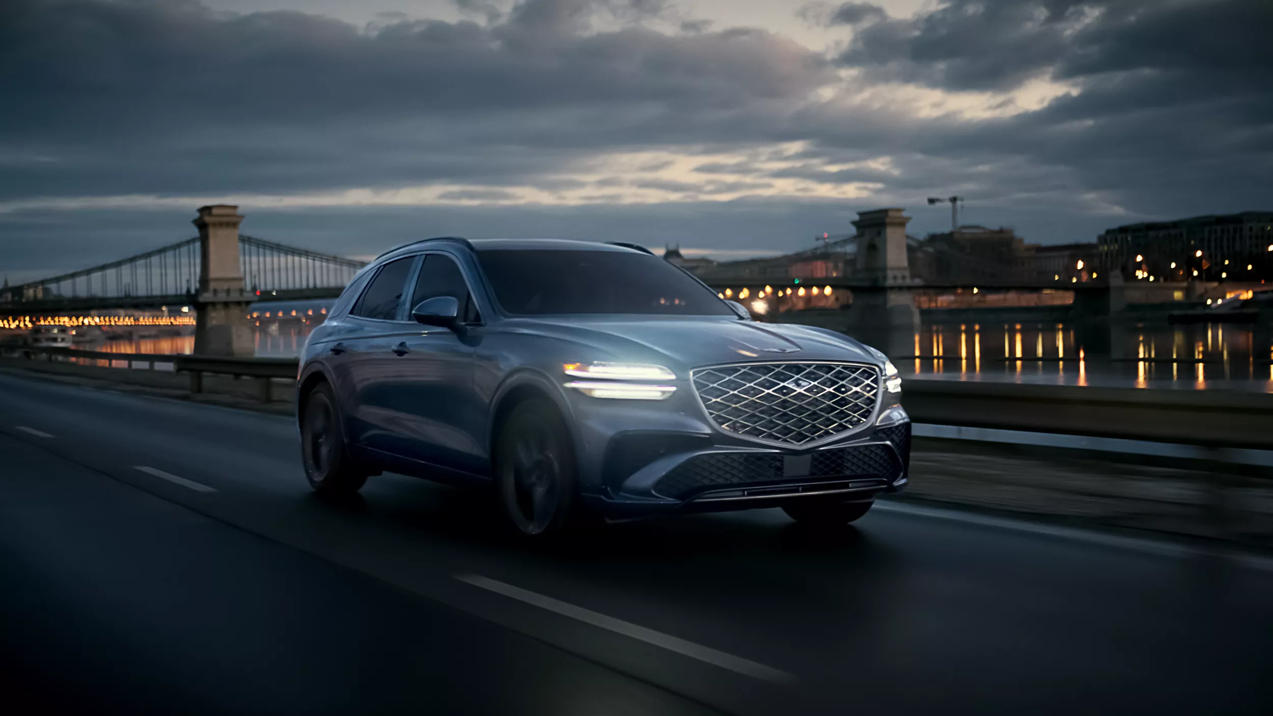 Front passenger side view of Genesis GV70 driving at night along a river with a city & bridge behind