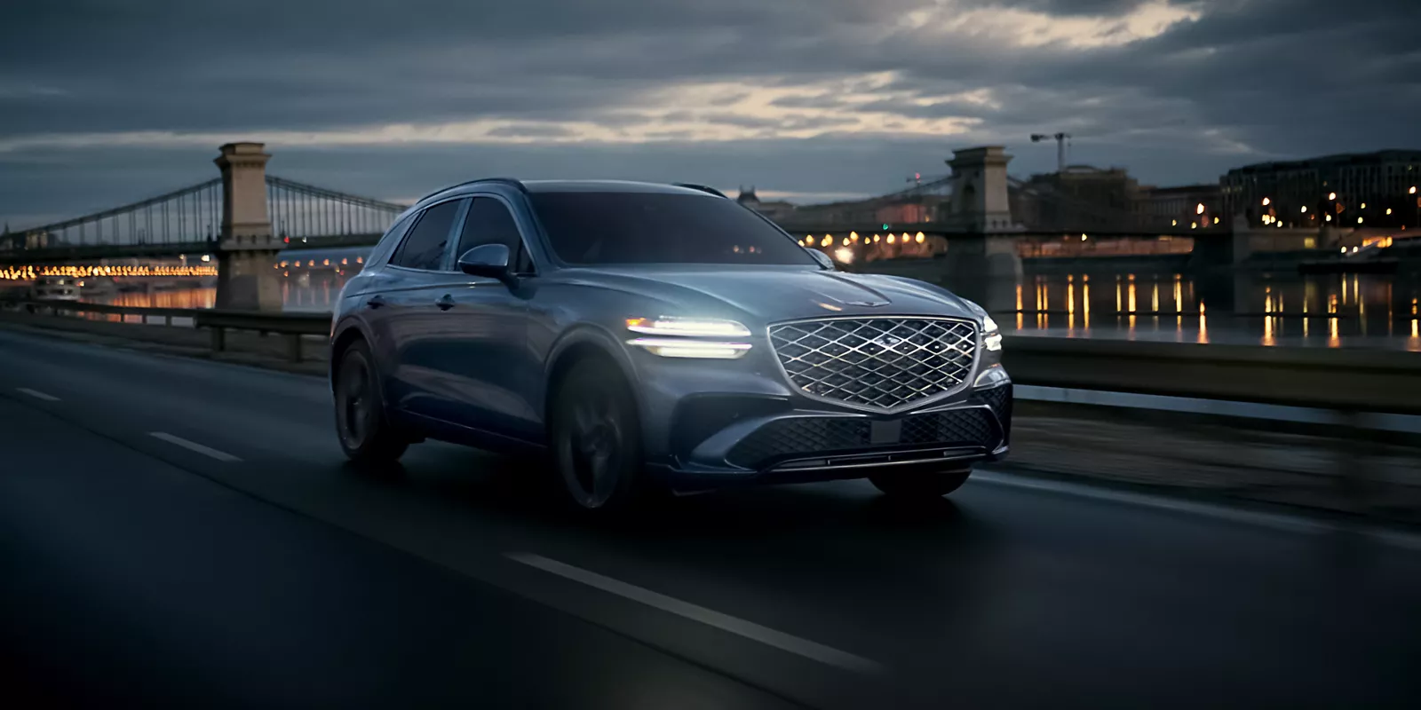 Front passenger side view of Genesis GV70 driving at night along a river with a city & bridge behind