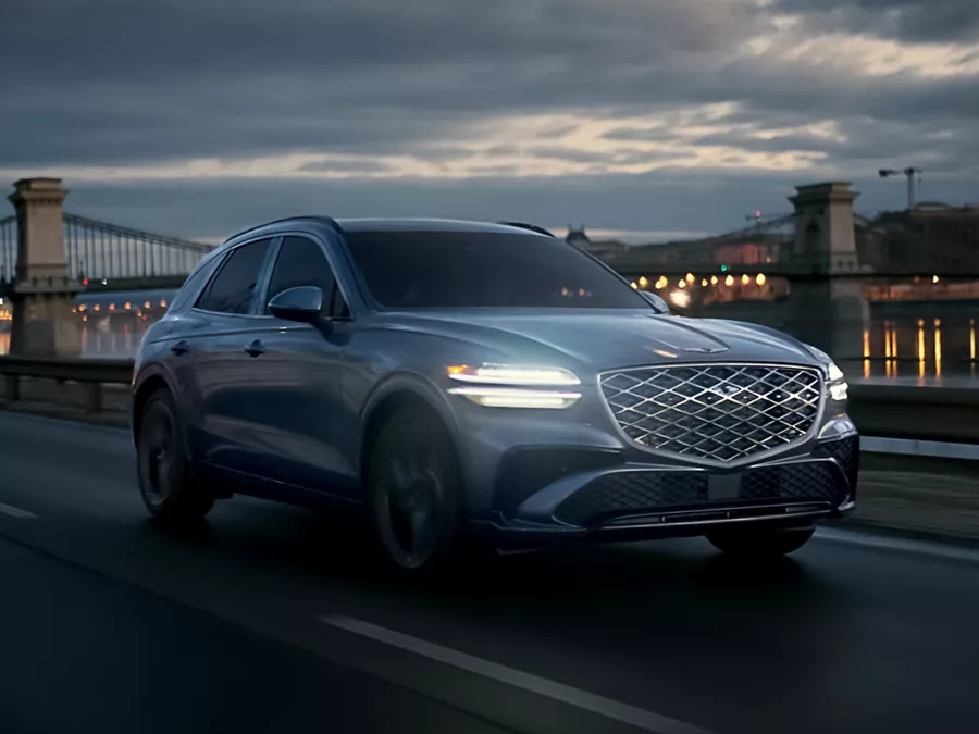 Front passenger side view of Genesis GV70 driving at night along a river with a city & bridge behind