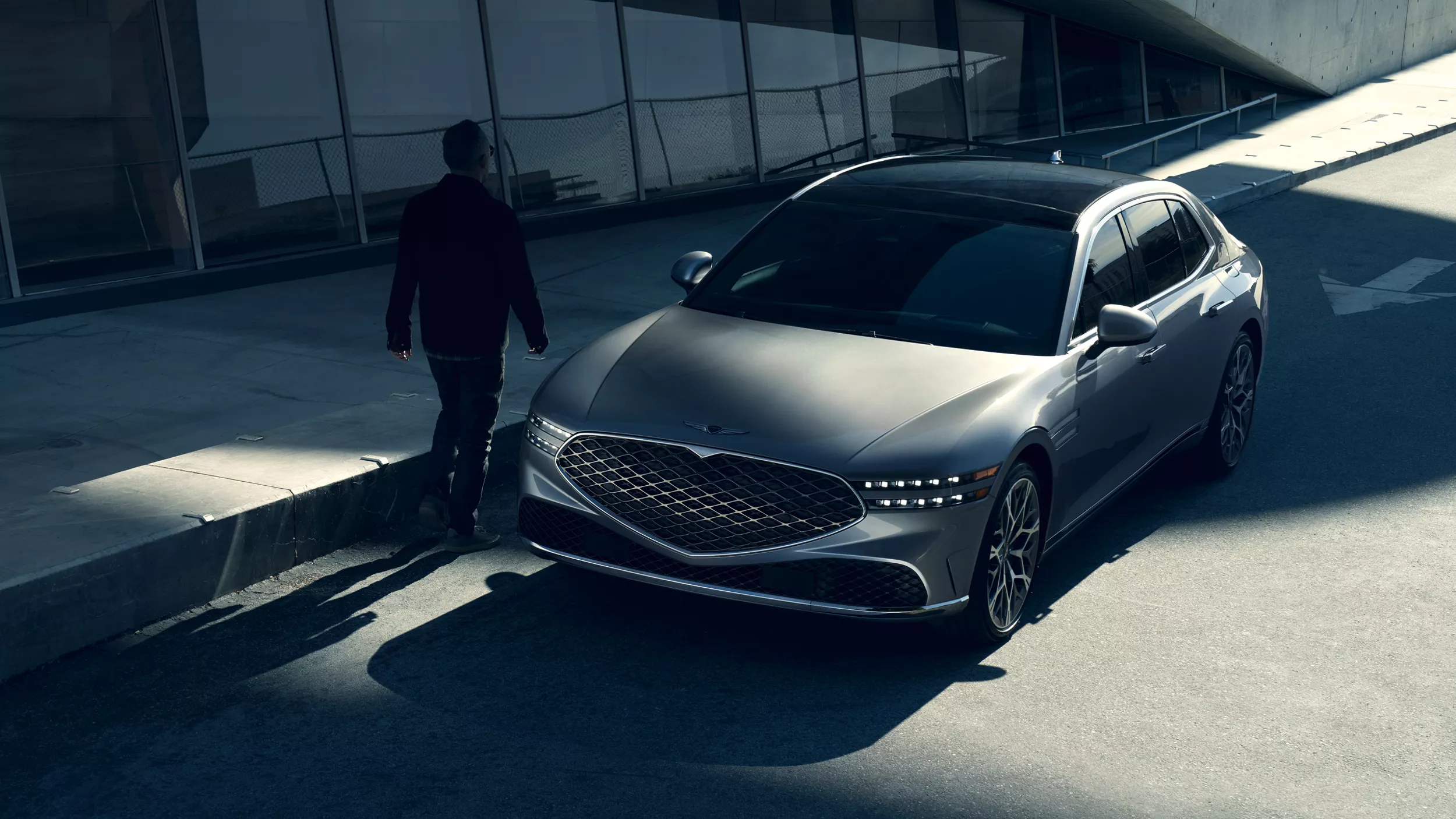 Overhead 3/4 front view of 2026 Genesis G90 in Savile Silver on a city street as person walks past