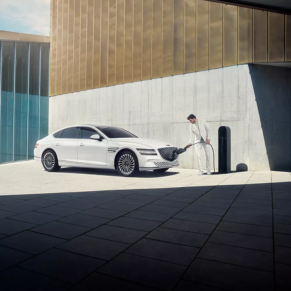 陽光下的白色 Electified G80 停在一棟現代化大樓旁，車影映在牆上。