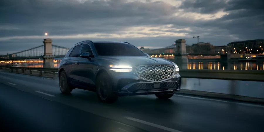 Front passenger side view of Genesis GV70 driving at night along a river with a city & bride behind