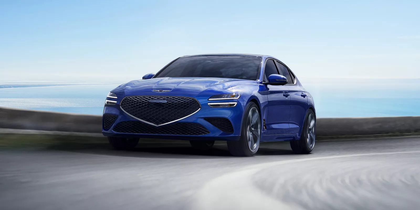 Front driver side action shot of Genesis G70 in Kawah Blue driving on a coastal highway