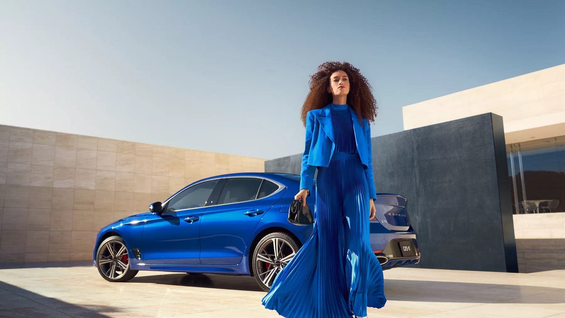 Genesis G70 (Kawah Blue) 停在頂層上有一女子走在車前的駕駛側視角照