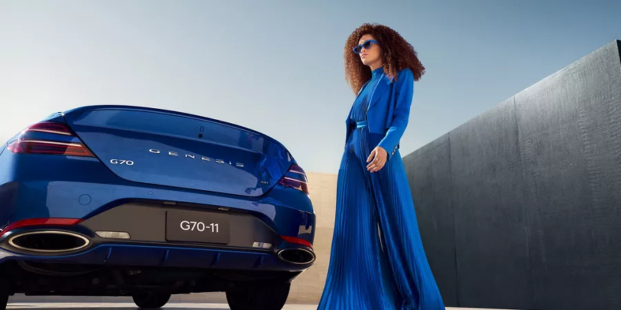 一藍衣女子走過停放的 Genesis G70 (Kawah Blue) 後方時所見後視照