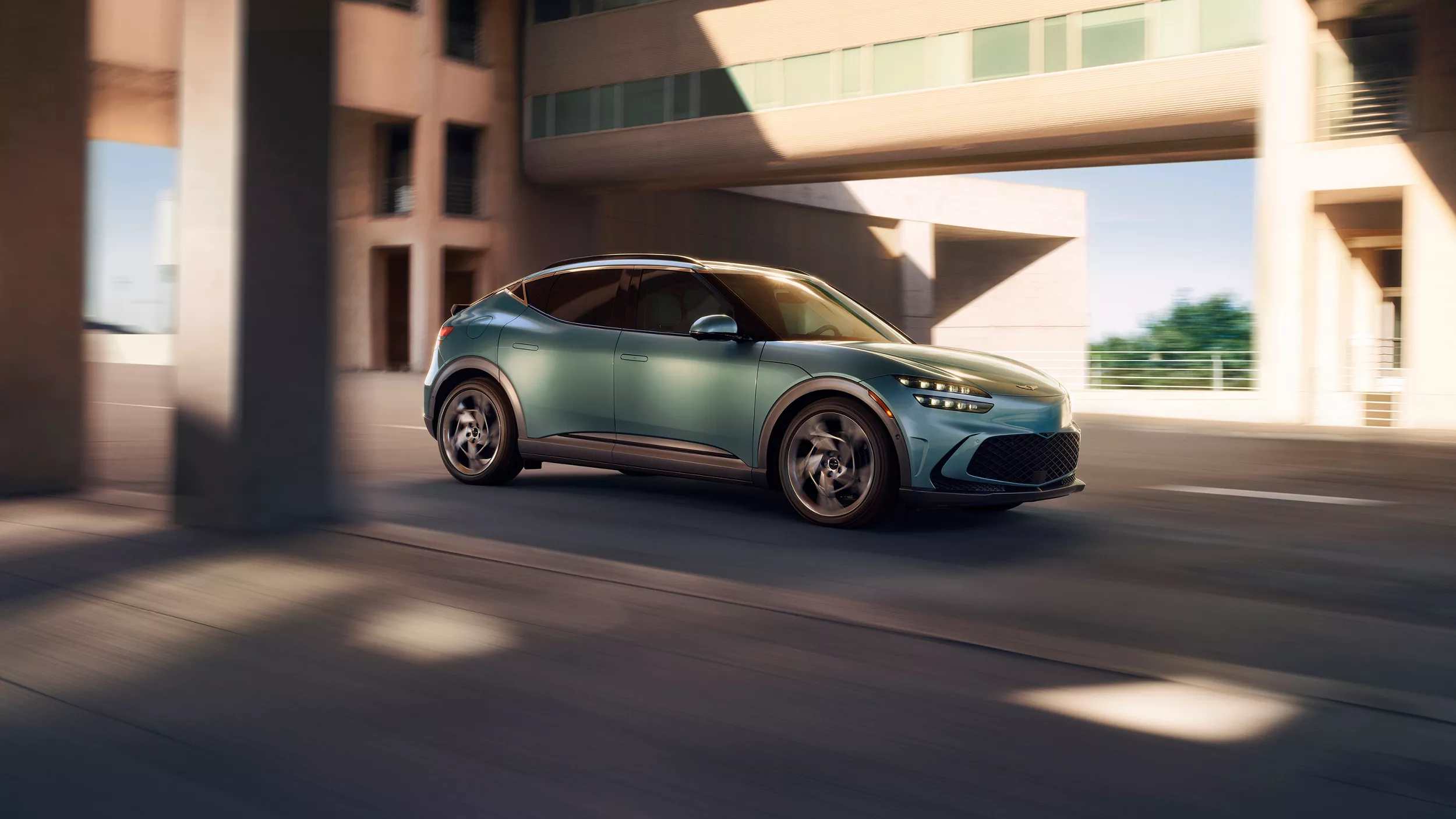 Passenger's side view of Genesis GV60 in Hanuama mint driving under modern building overhang