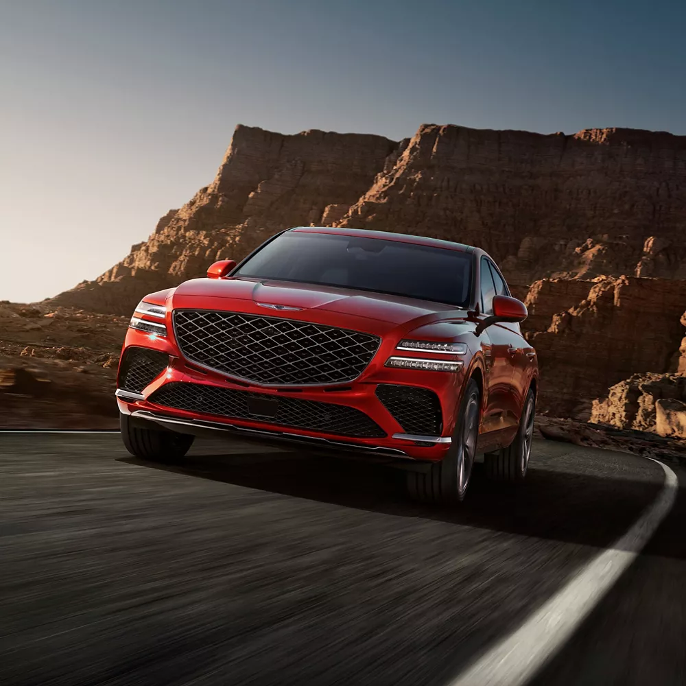 3/4 front driver side view of Genesis GV80 Coupe in Mauna Red driving along desert mountain at dusk