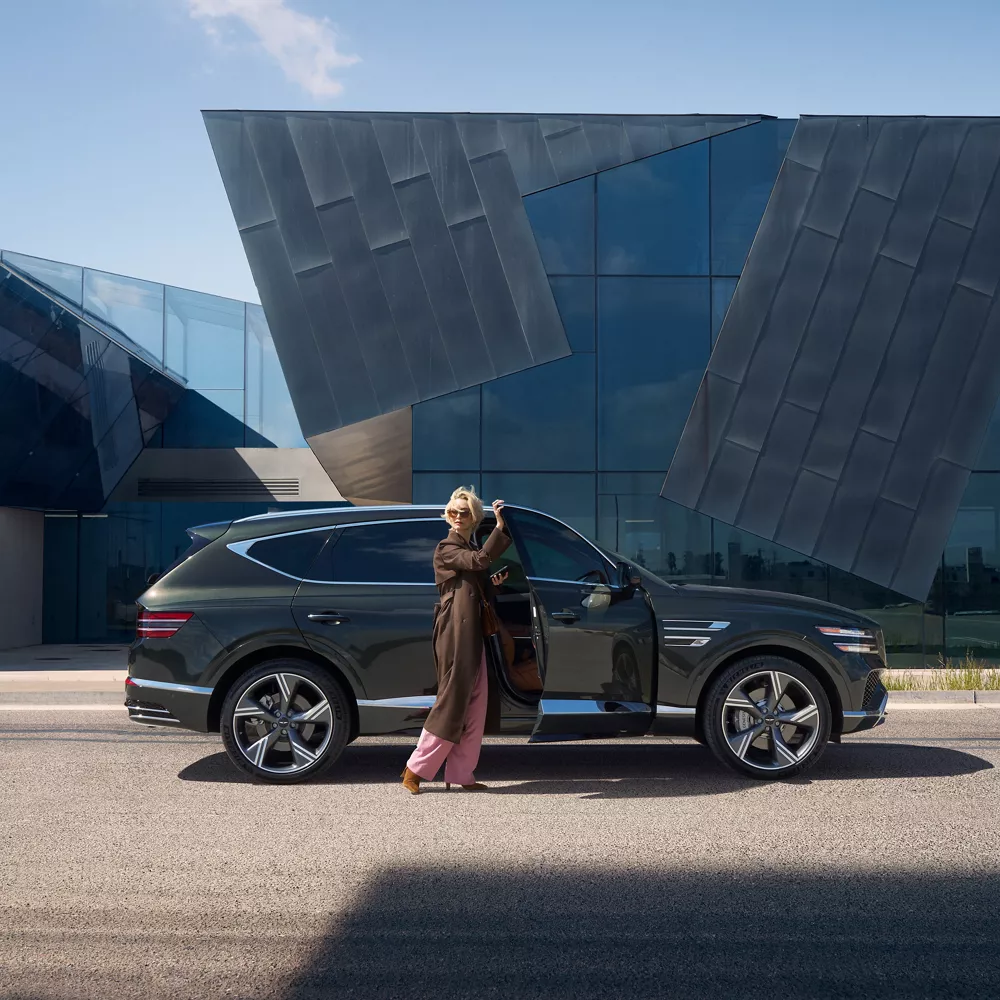 Passenger side profile of Genesis GV80 parked by modern building with driver opening passenger door
