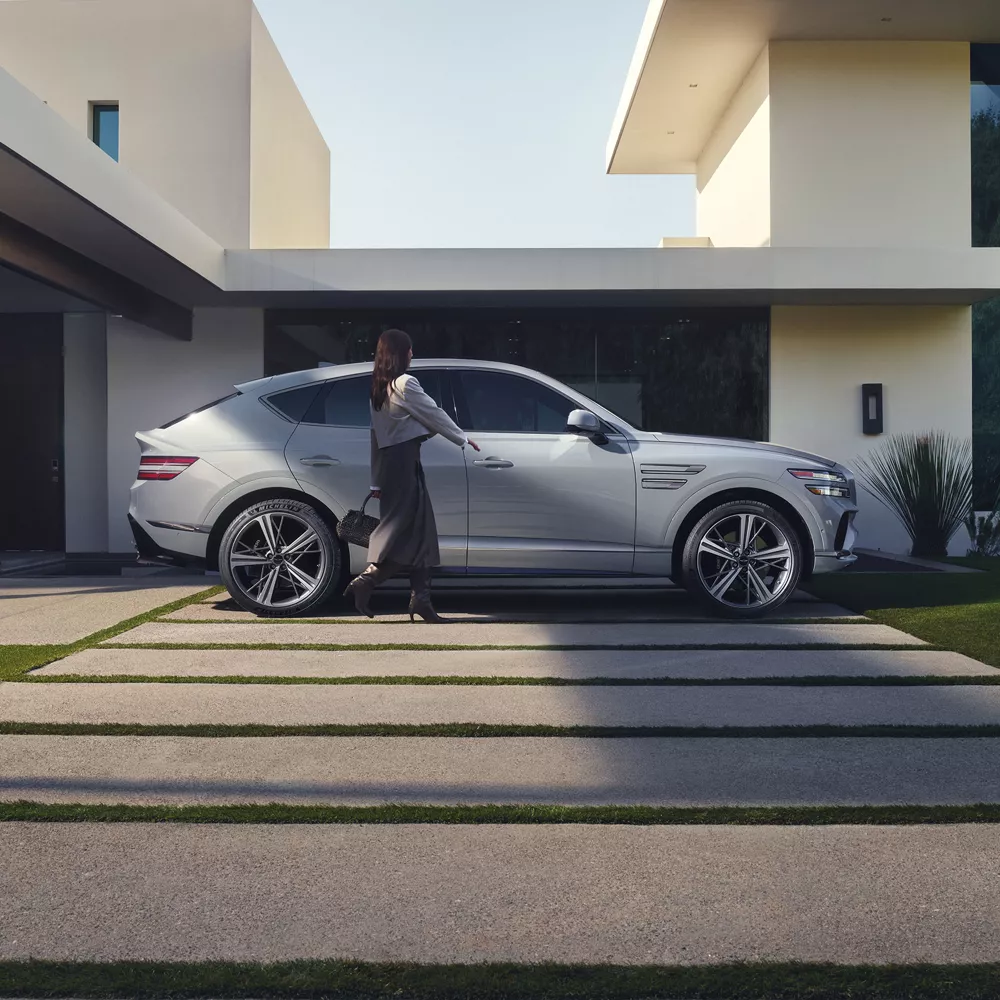 Passenger side profile view of Genesis GV80 Coupe in Bering Blue parked at modern home as couple walks to vehicle