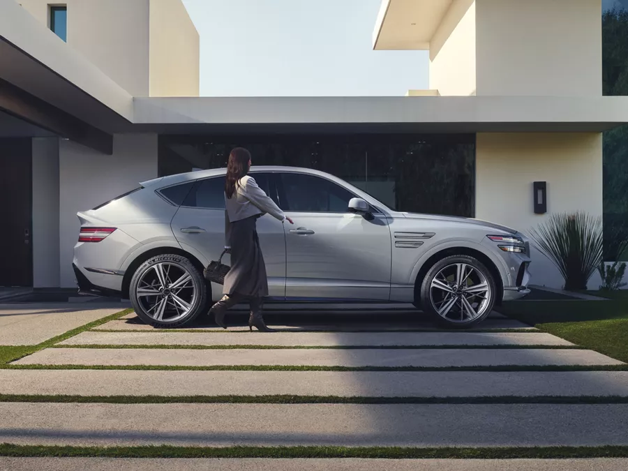 Passenger side profile view of Genesis GV80 Coupe in Bering Blue parked at modern home as couple walks to vehicle