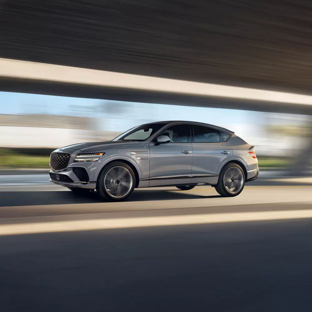 Driver side profile action shot of Genesis GV80 Coupe in Bering Blue driving through bridge/tunnel