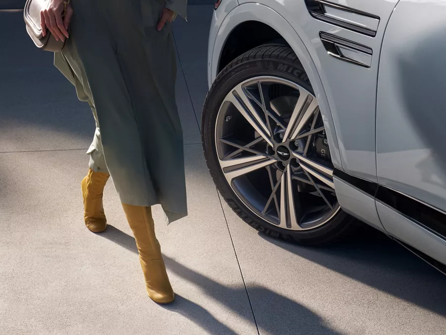 Close up of 20-inch dark gray matte rims on Genesis GV80 Coupe in Bering Blue, woman in dress nearby