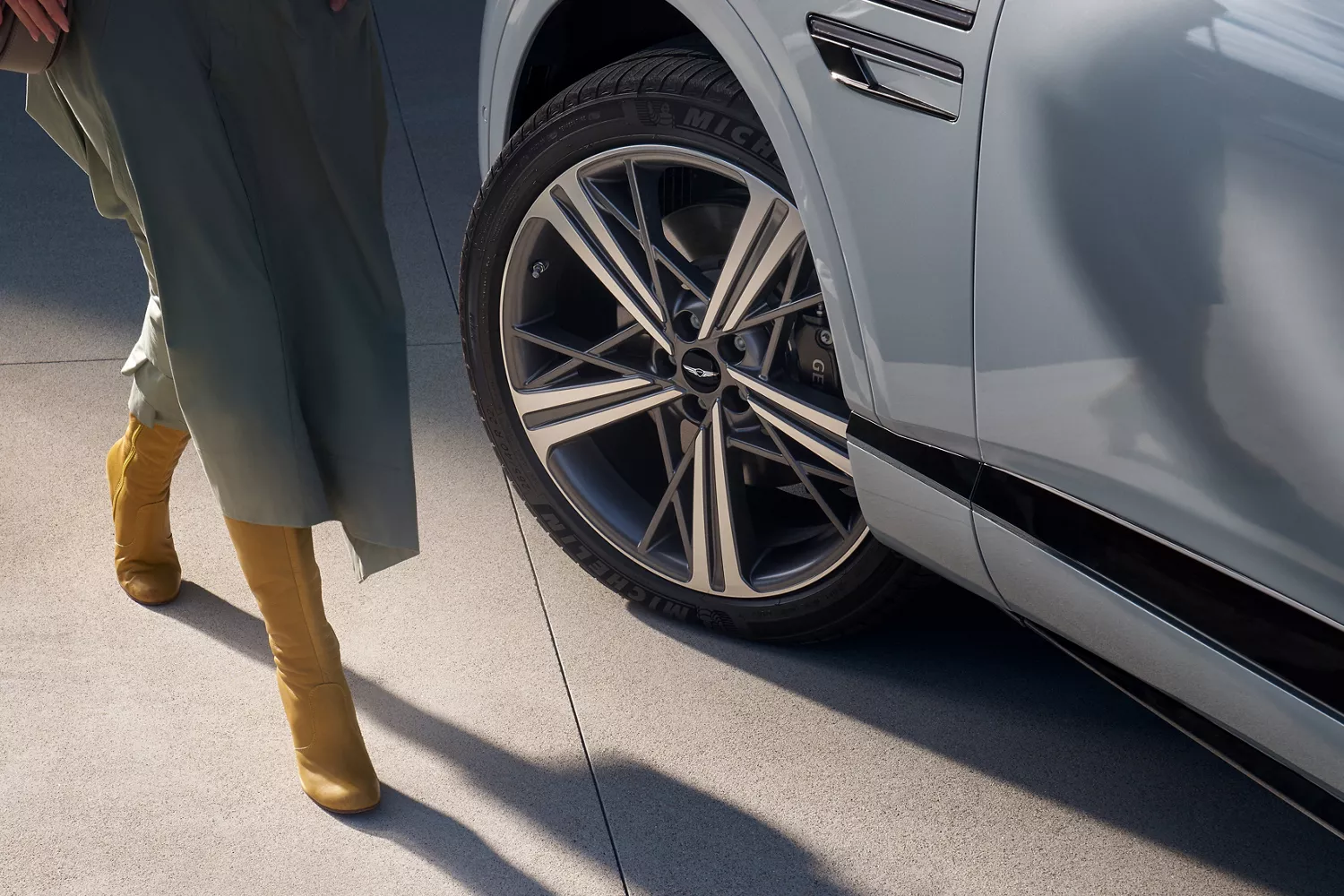 Close up of 20-inch dark gray matte rims on Genesis GV80 Coupe in Bering Blue, woman in dress nearby