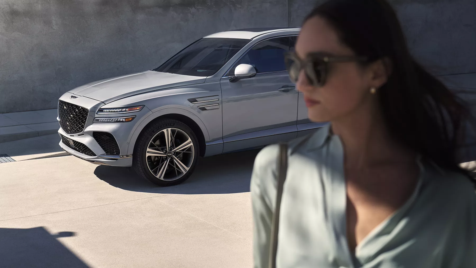 3/4 front driver side view of Genesis GV80 Coupe in Bering Blue parked near wall with woman in foreground