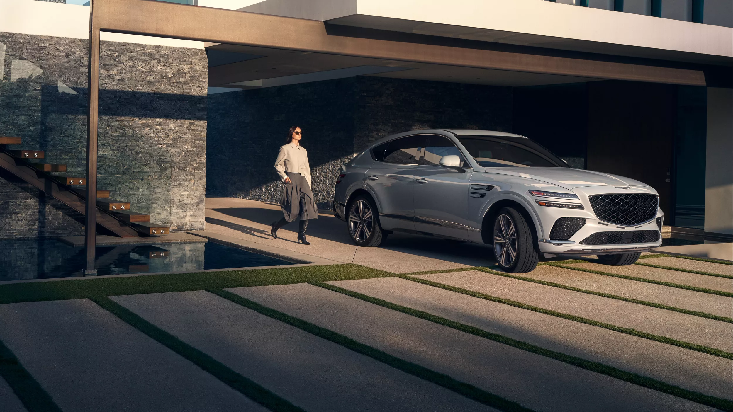 3/4 front passenger side view of woman walking beside Bering Blue Genesis GV80 Coupe at modern home