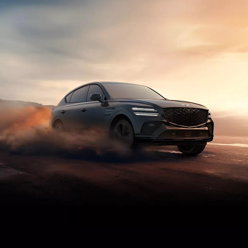 3/4 front passenger side view of Genesis GV80 Coupe driving on dusty road at sunset