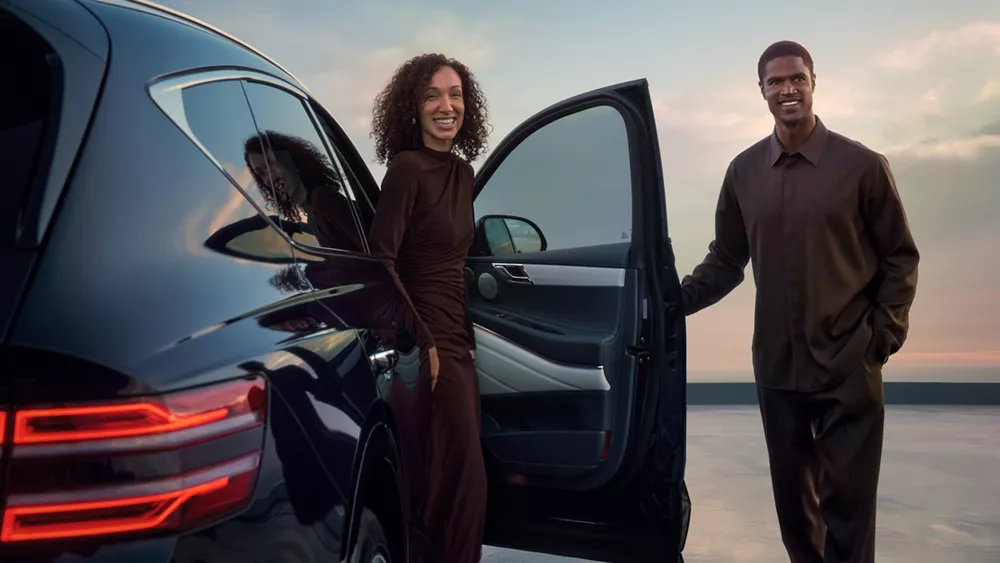 3/4 rear passenger view of two people dressed in brown standing beside Genesis GV80 in Capri Blue