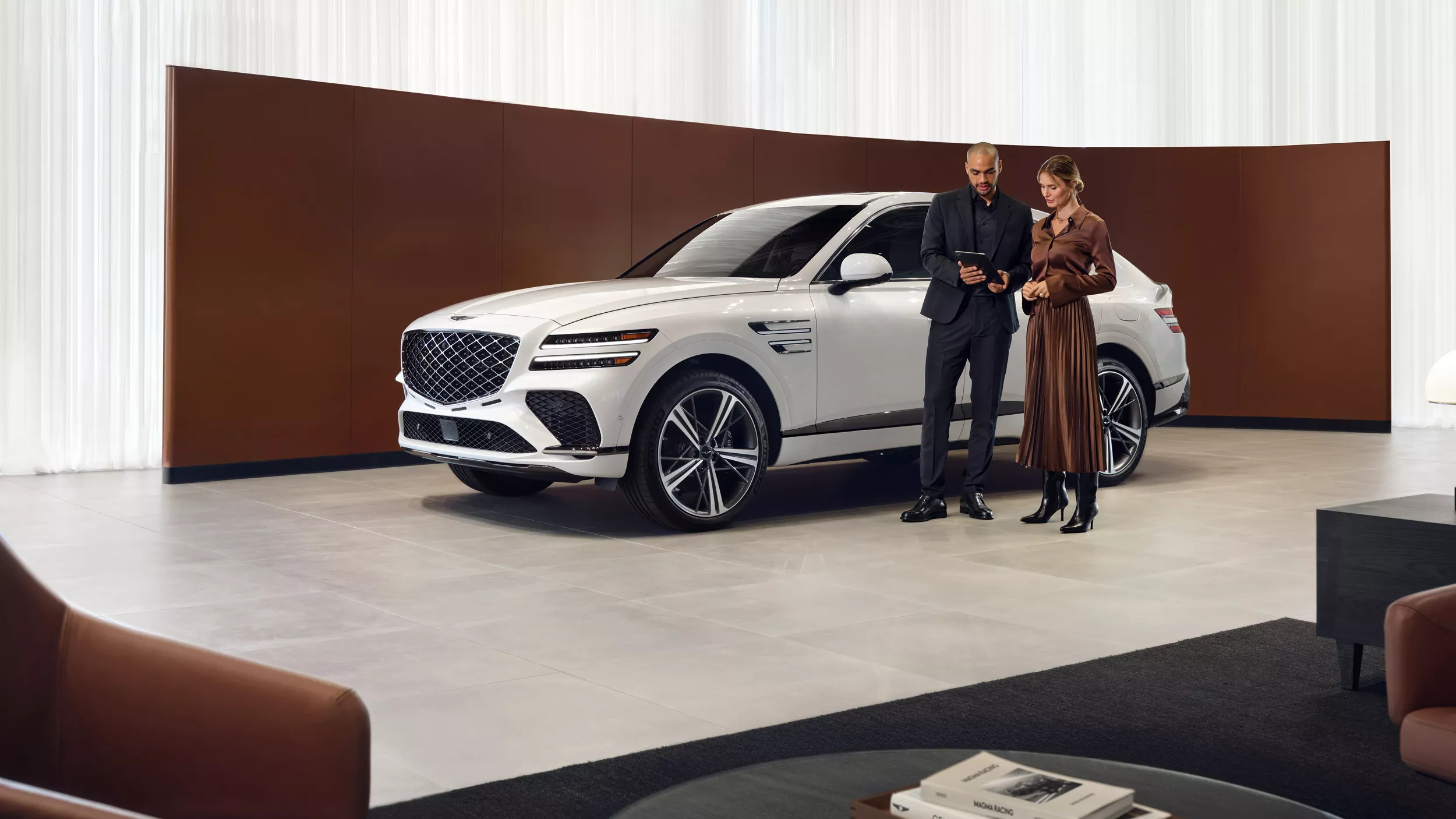 Two people standing in white & brown accented showroom, Genesis GV80 Coupe in Vatna Gray behind them