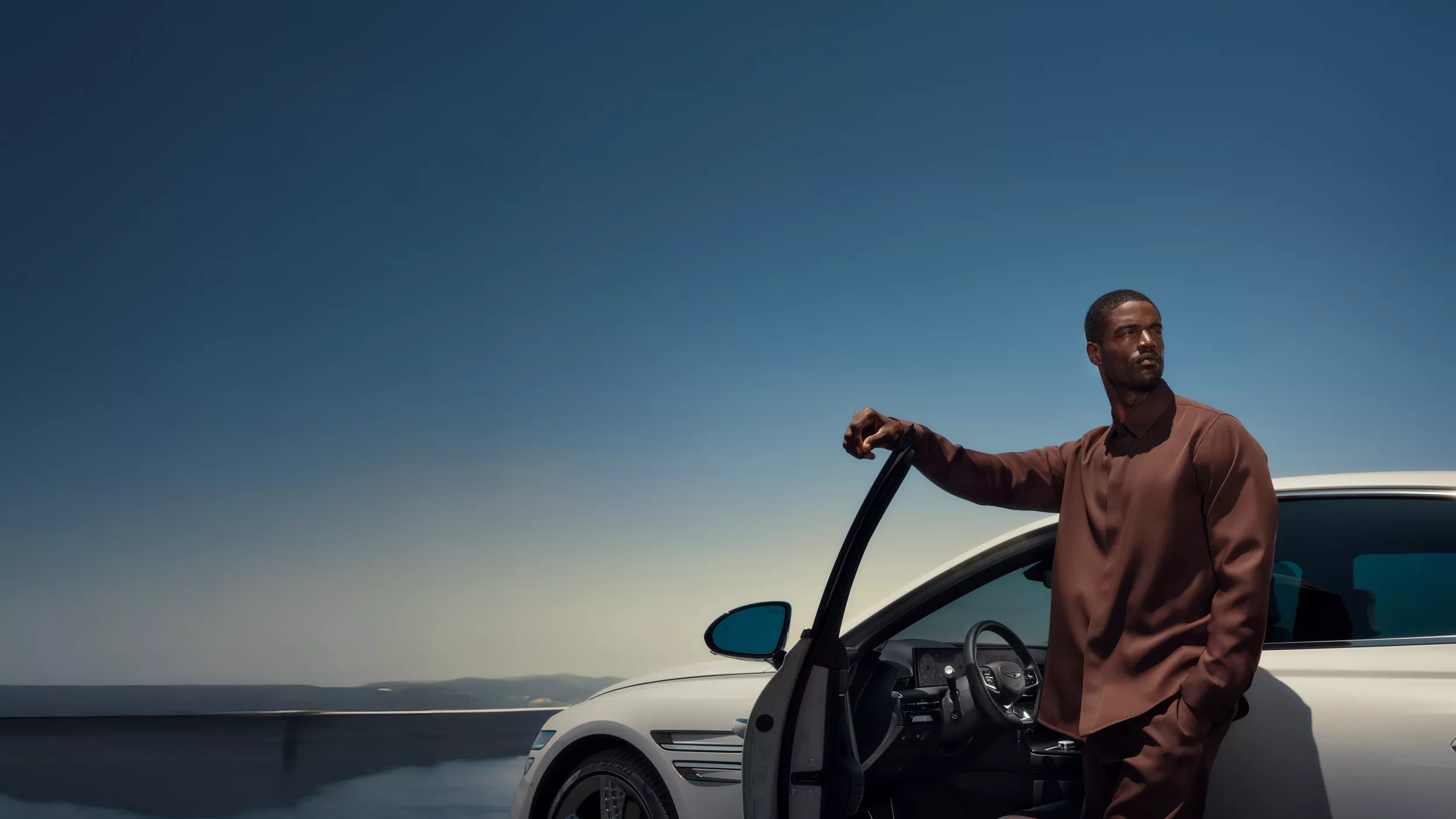 Man standing next to open driver side door of Genesis G80 in Vatna Gray, blue sky in background