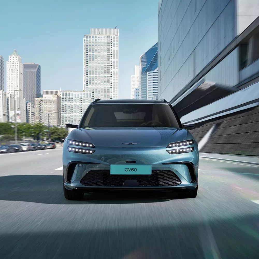 2026 Genesis GV60 in Tomso Green driving toward viewer on city road with skyline in background